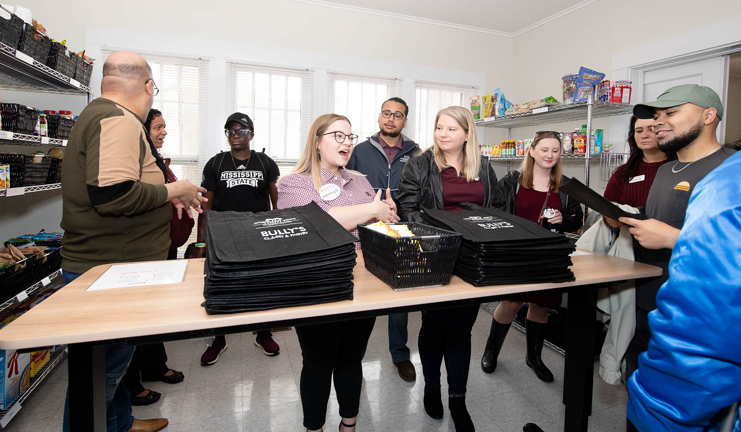 Female in maroon and white checkerboard shirt talking to group surrounding table with cloth shopping bags and pantry items in background