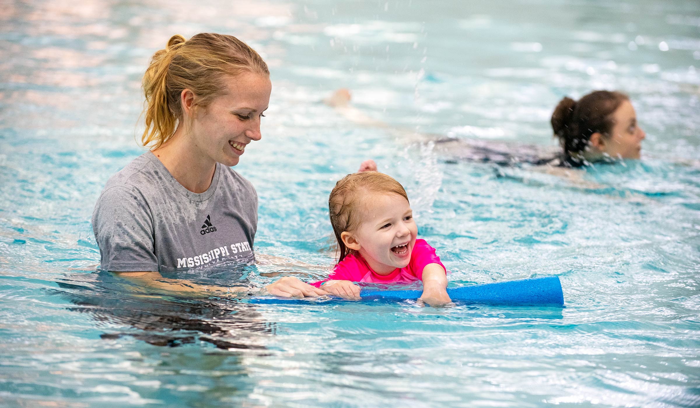 Swim instructor in gray shirt in swimming pool with child in pink bathing suit practicing kicking with blue pool noodle