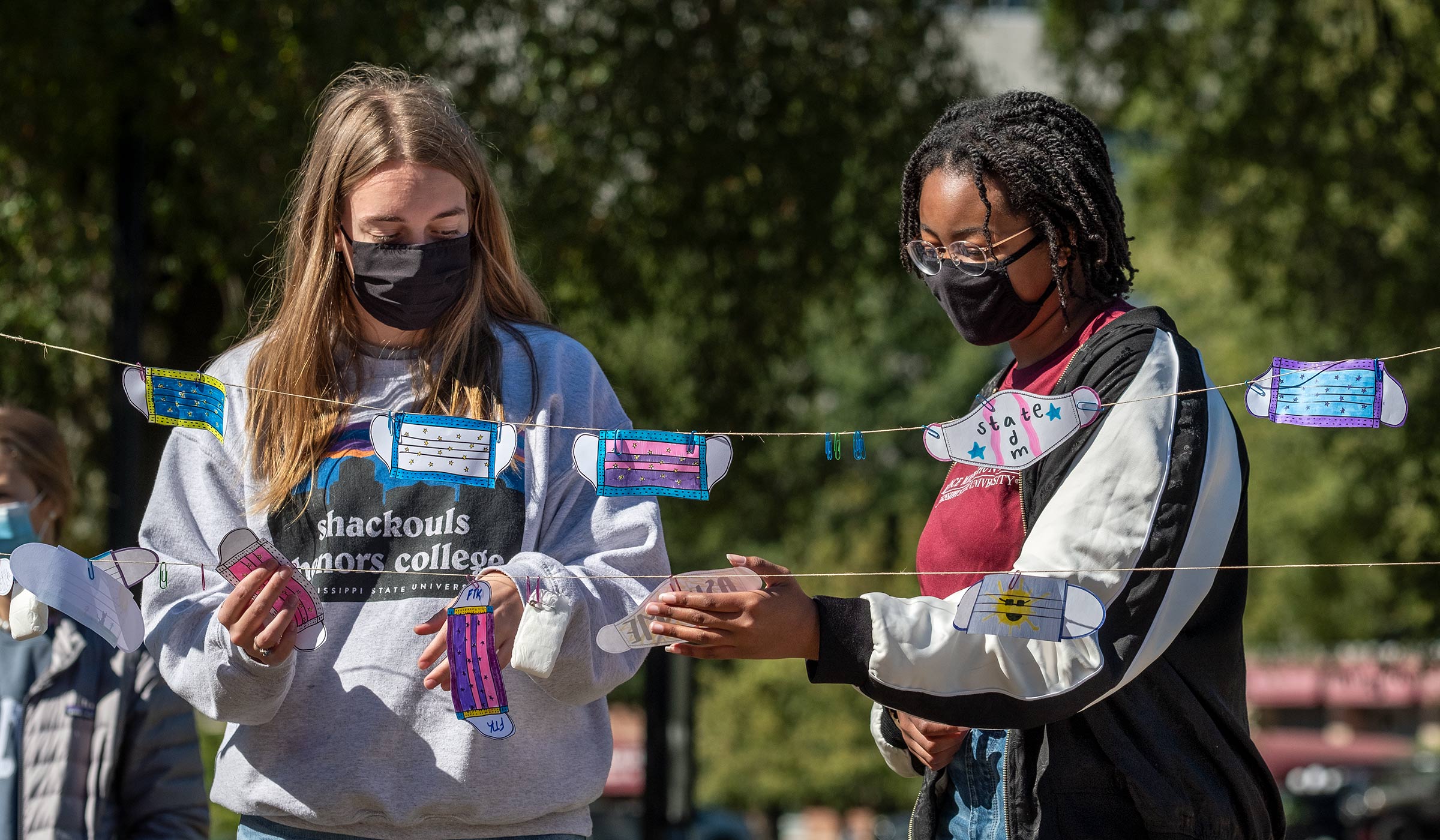 Two Dance Marathon representatives arrange decorated paper mask cutouts clipped to a line to promote Dance Marathon.