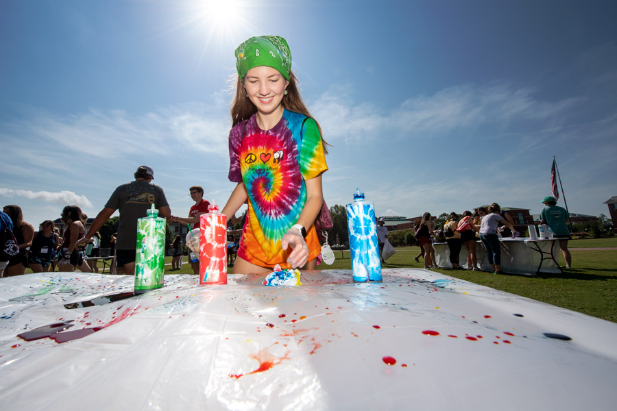 Wearing a tie-dye shirt and green bandana, a female student does more tie-dye at a Drill Field table.