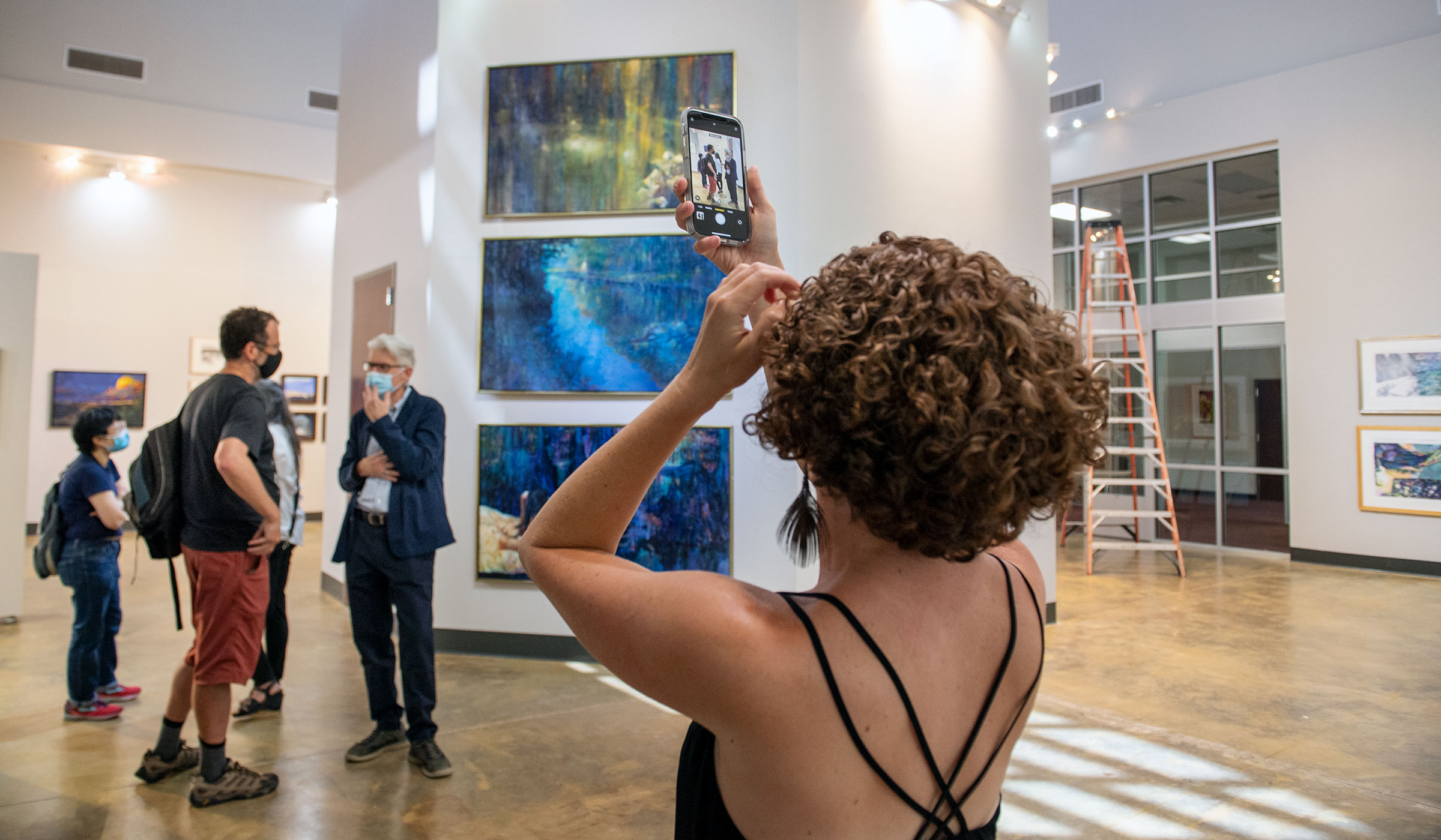 Art Professor Emeritus Brent Funderburk speaks with current art professors (all wearing facemasks) in the Welcome Center art gallery, with the back of the Exhibition Coordinator, Dixie Boswell, in the foreground taking a cell phone picture.