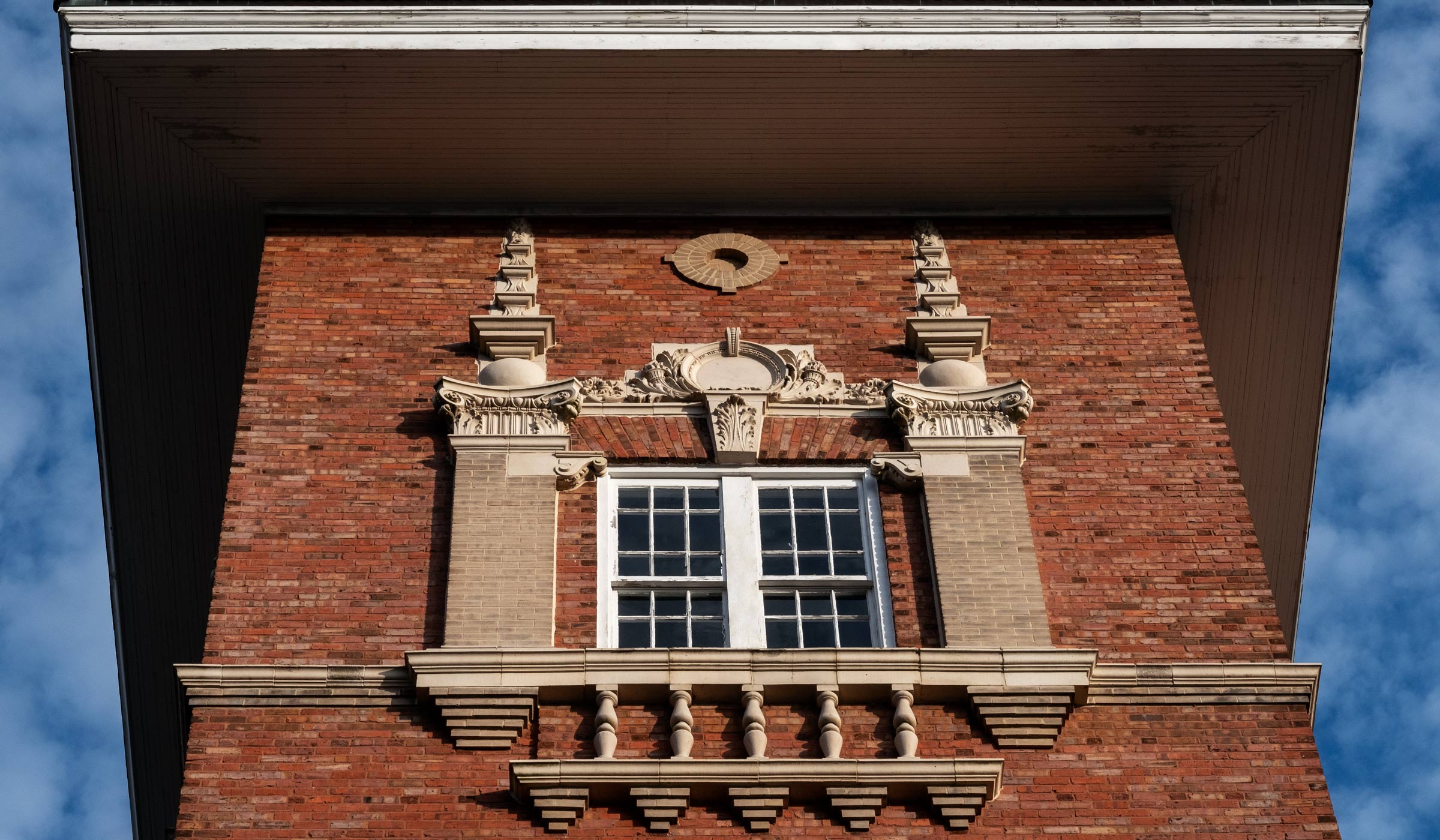 The tops of each of the two red brick towers of the historic Industrial Education building are adorned with detailed craftsmanship. The photo focuses on the top of one tower, with lightly clouded blue sky in the background.