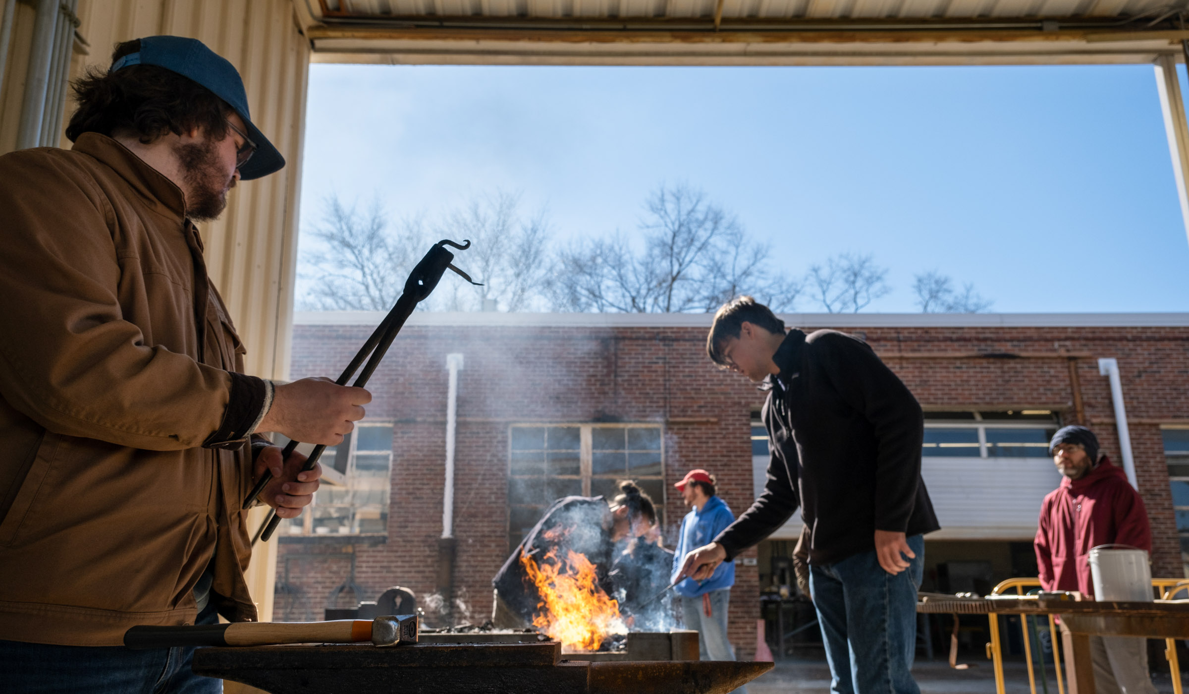 A workshop participant holds a completed iron hook up in the foreground while others work at the fire and a second anvil in the background, with blue sky beyond.