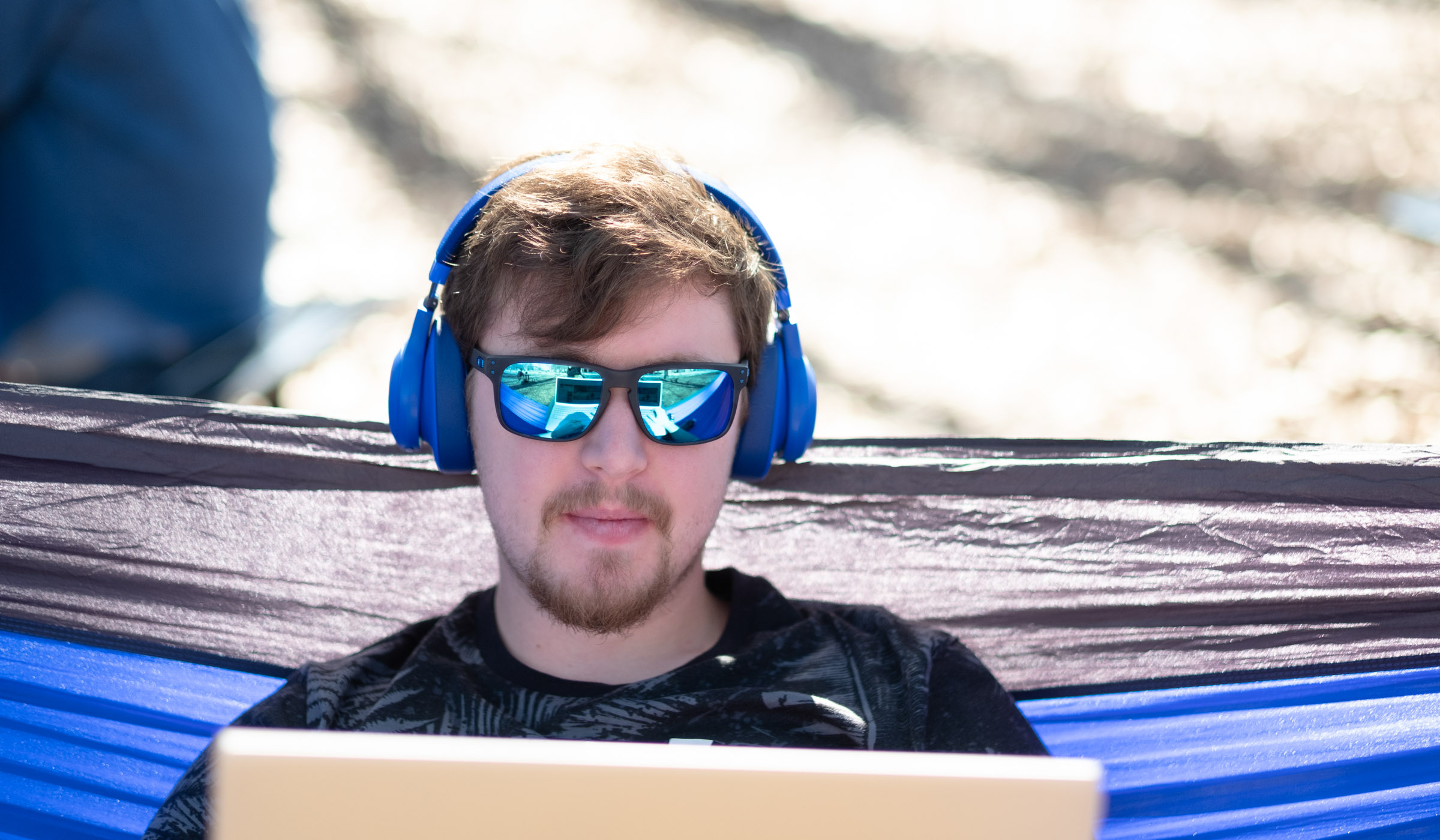 A tight crop on the face of a male student sitting in a blue hamock and wearing sunglasses with blue lenses, reflecting the laptop in his lap.