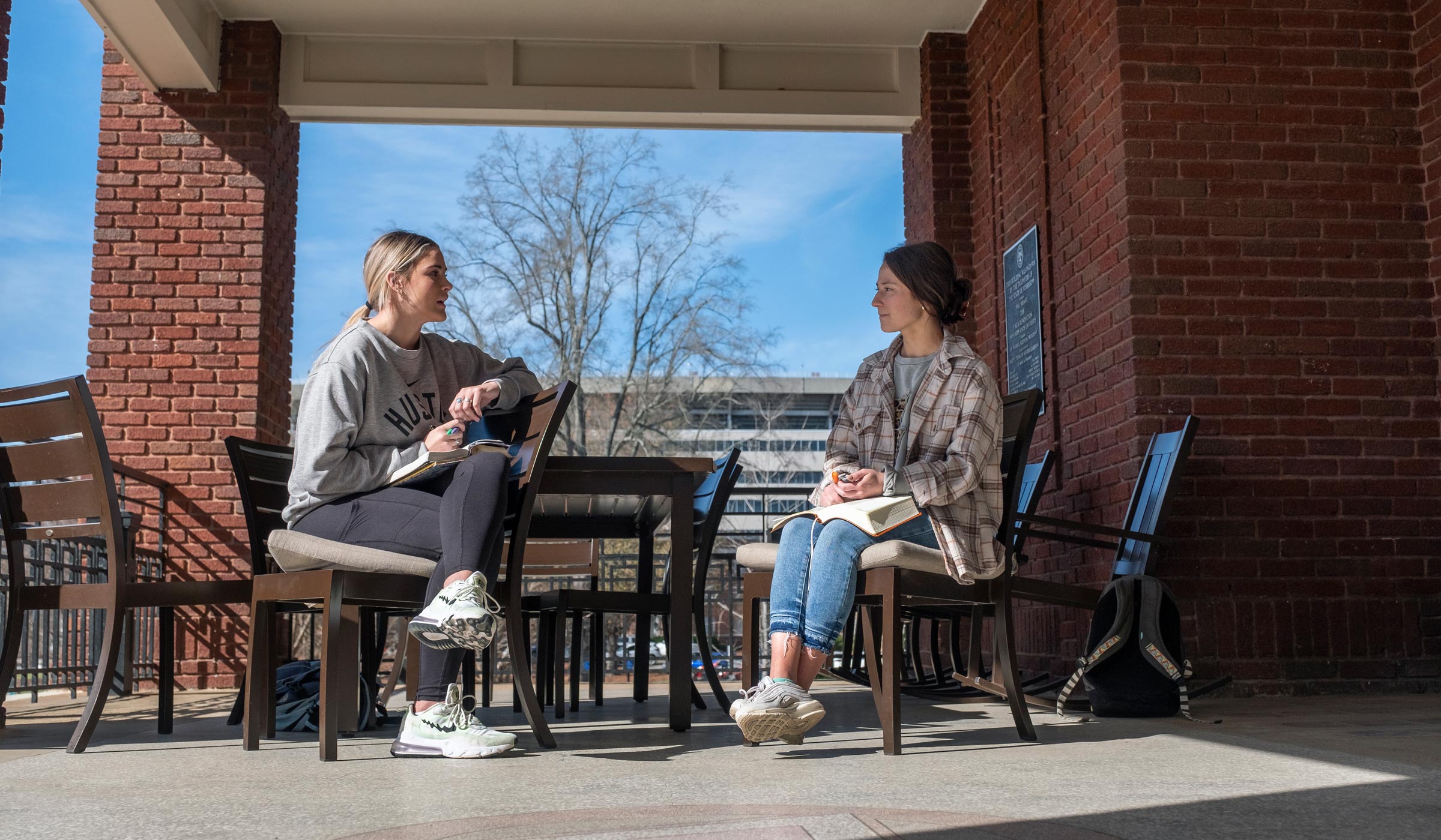 Two students sit and visit at a table on the front porch of the YMCA building, with blue sky showing beyond.