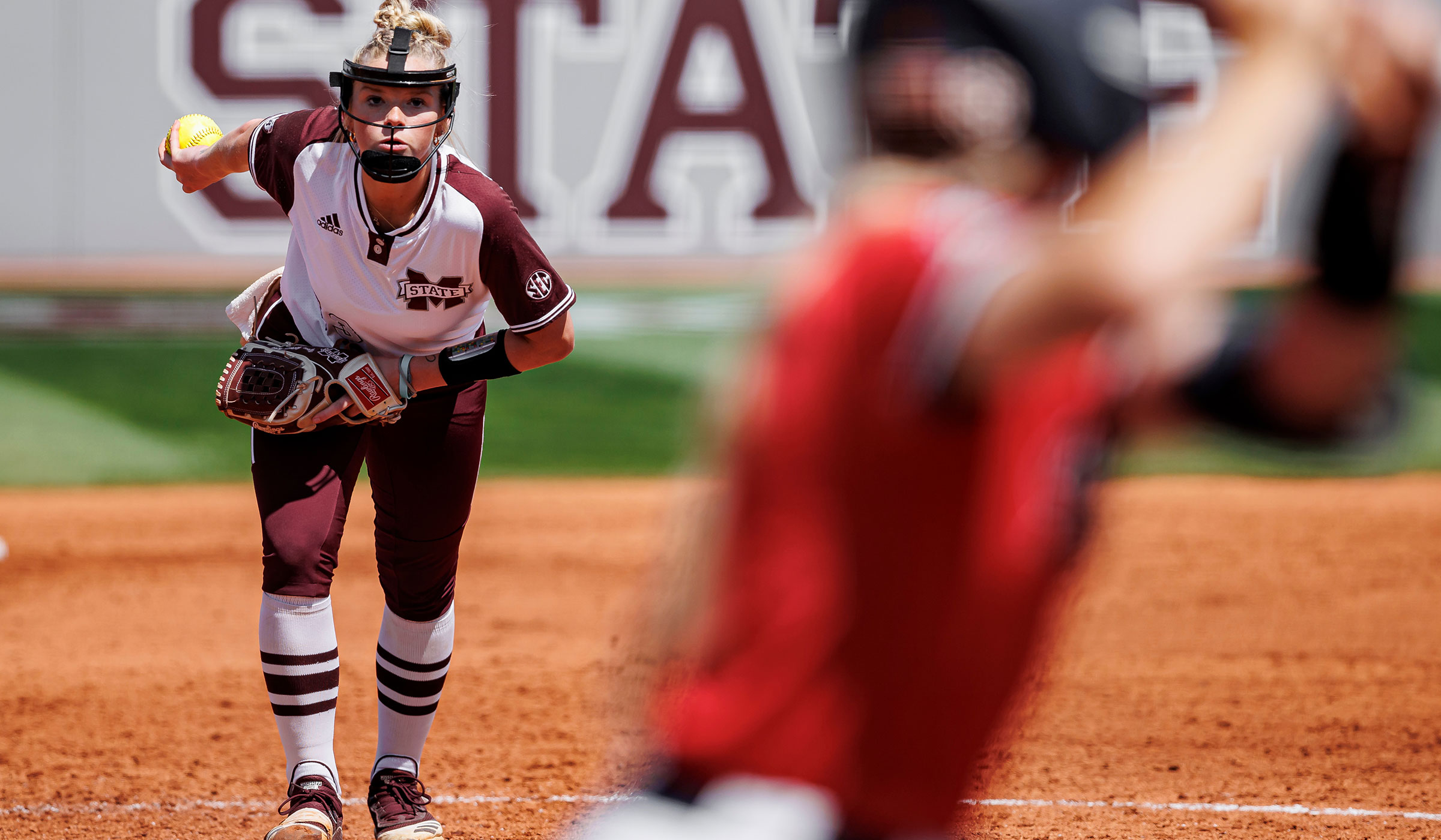 Female in maroon and white uniform winding up to pitch a neon softball to batter at plate wearing red.