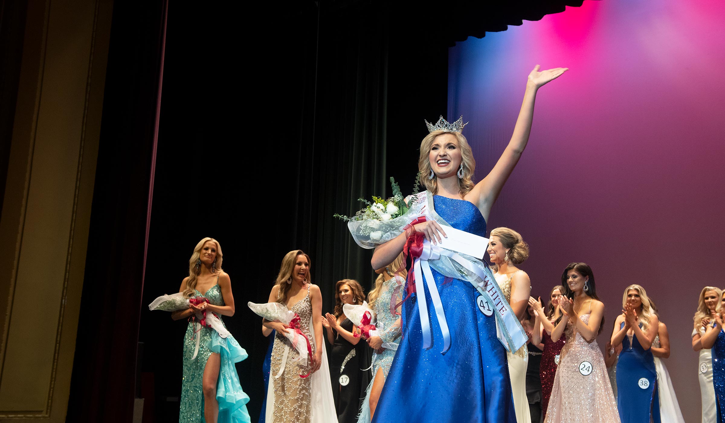 Female in blue gown with sash, crown, and flowers waving at crowd