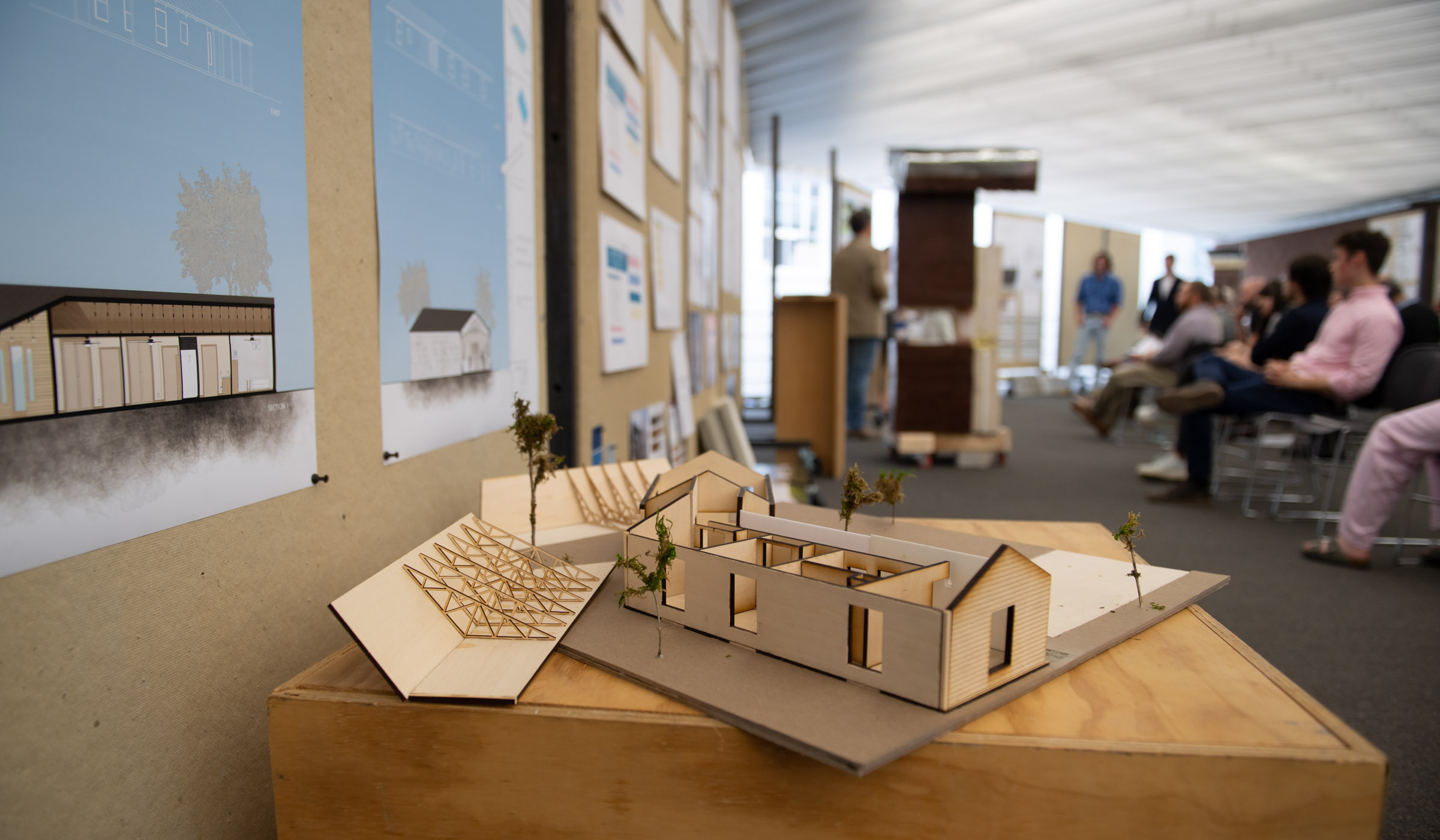 A 3D architectural model of a house is in focus in the foreground on a wooden pedestal, with students presenting their projects to seated attendees in the background.