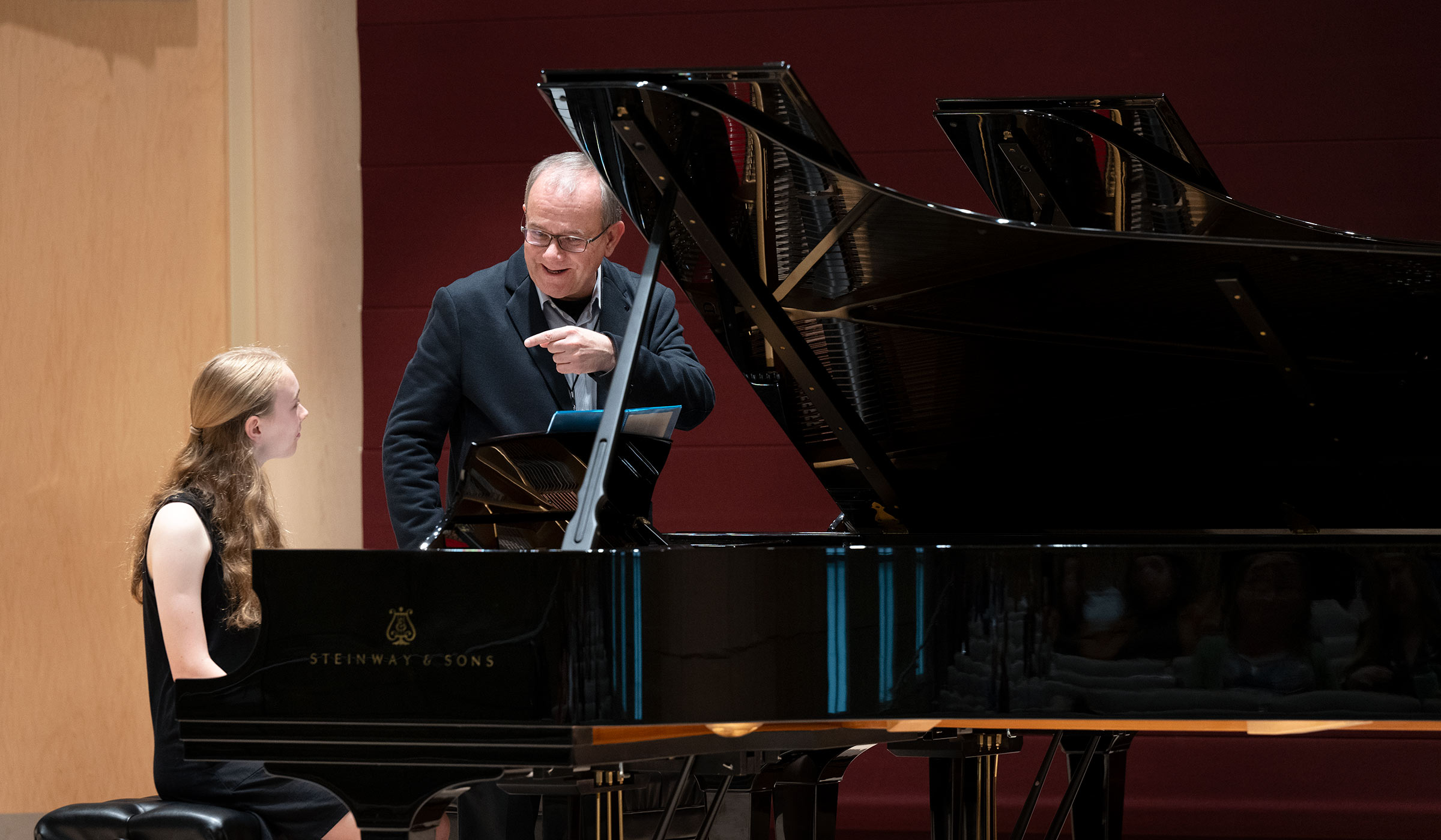 Man in jacket gesturing near piano to female in black dress