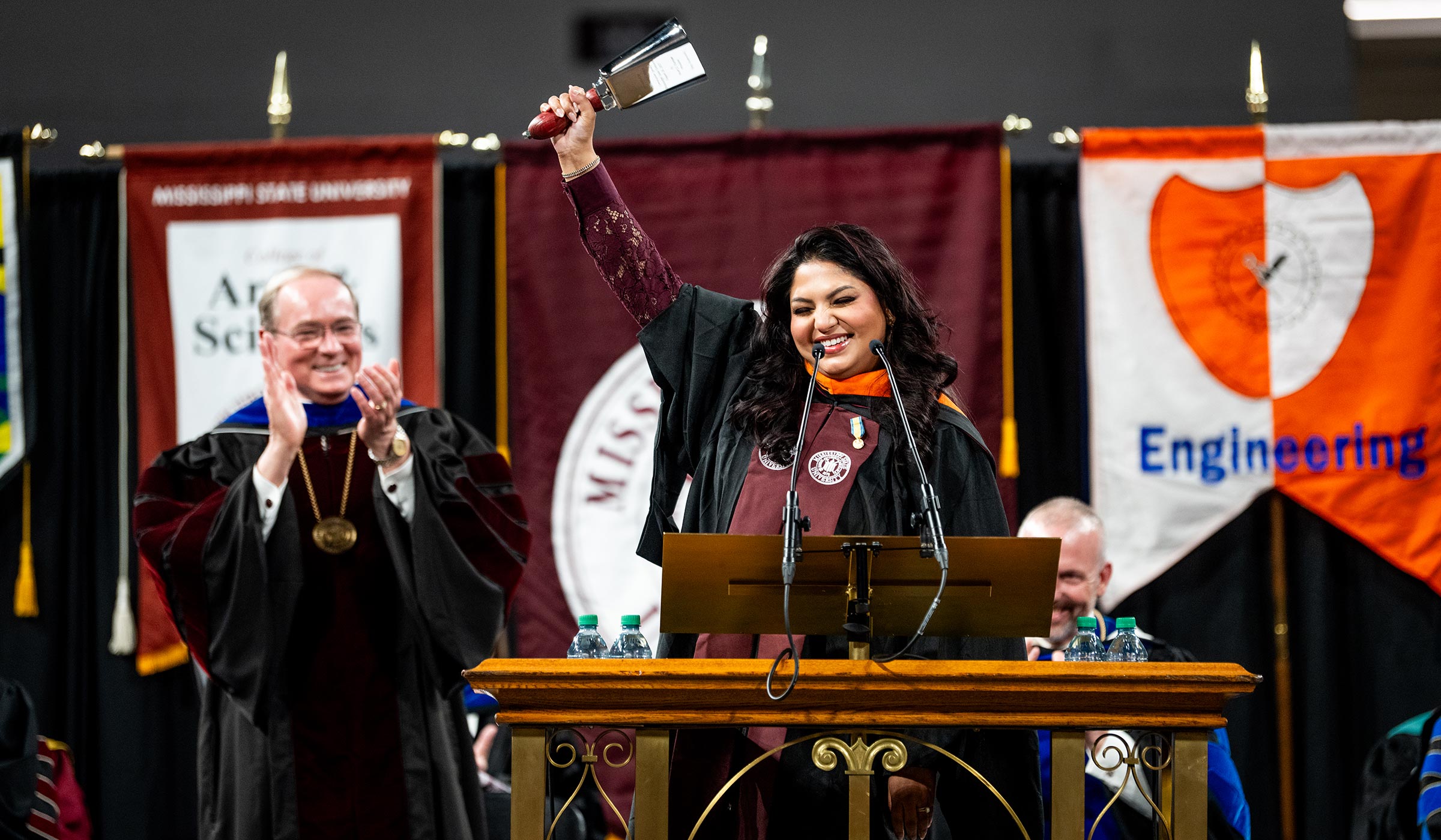 Woman in robed regalia ringing cowbell with colorful flags behind her