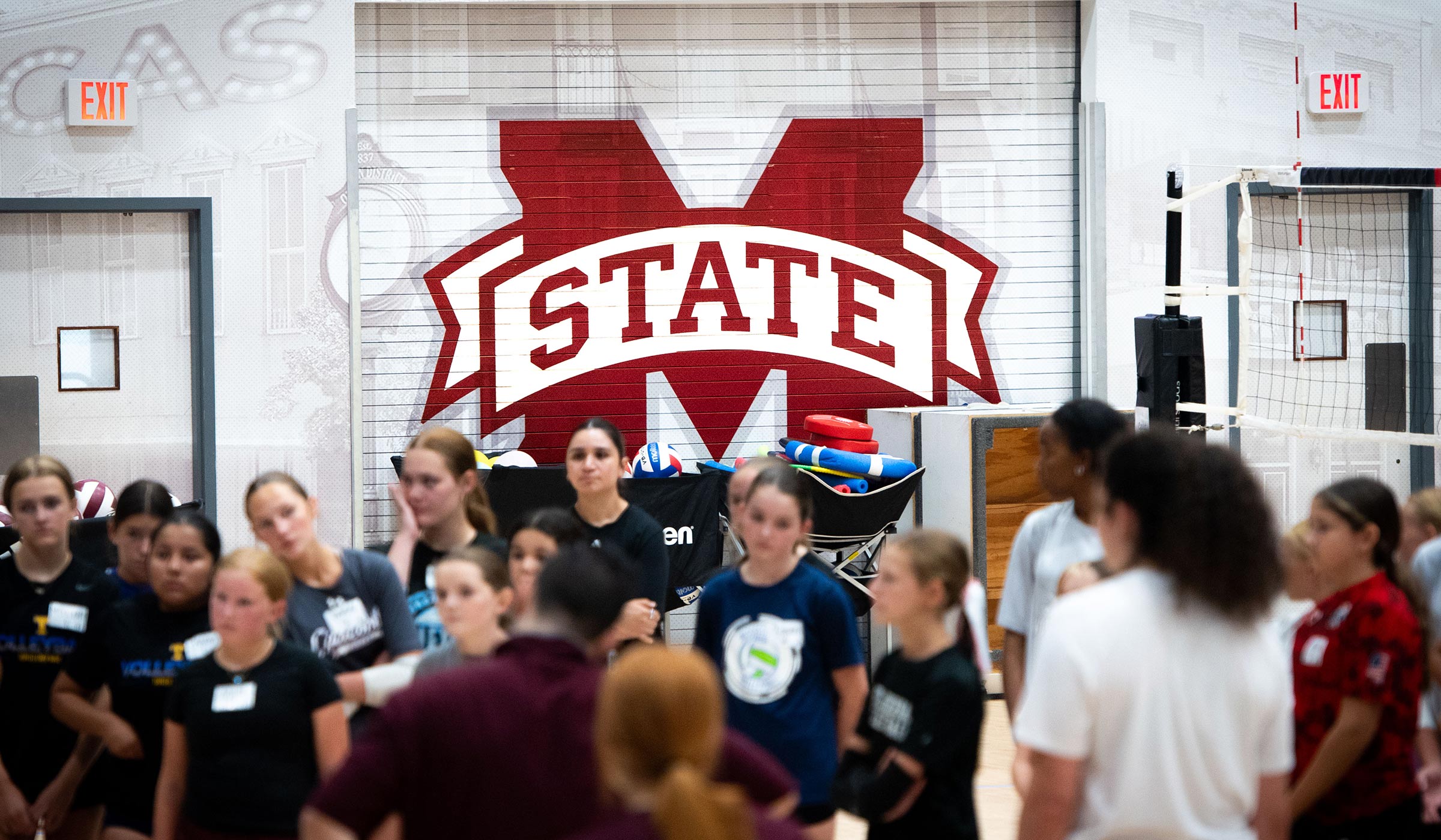 MSU volleyabll camp participants gather on the gym floor in front of a large MState logo displayed on the wall.