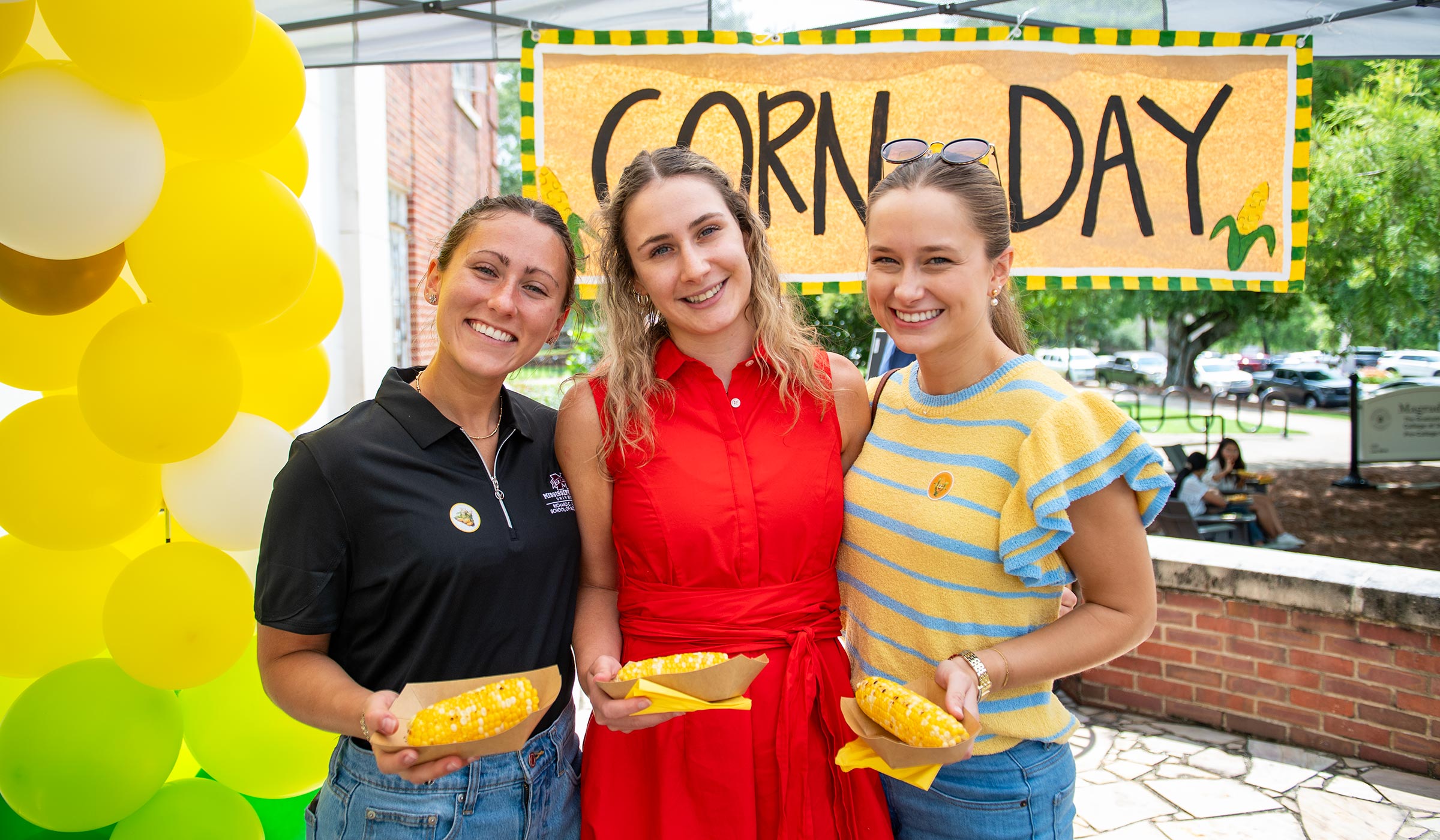 Students pose with corn in hand in front of a banner decorated for Corn Day.