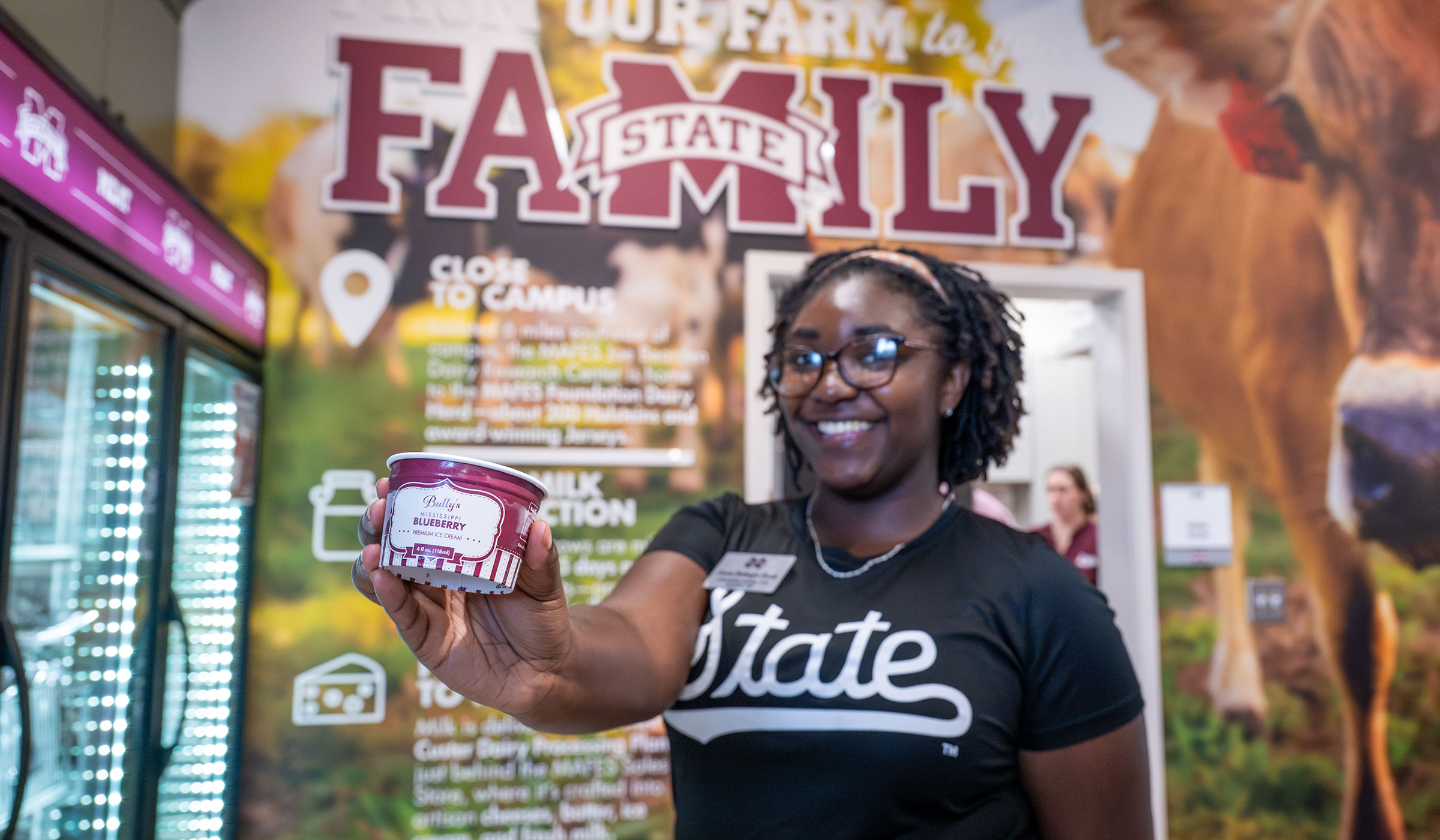 A student poses with blueberry ice cream in hand in front of a decorative wall in the MAFES store.