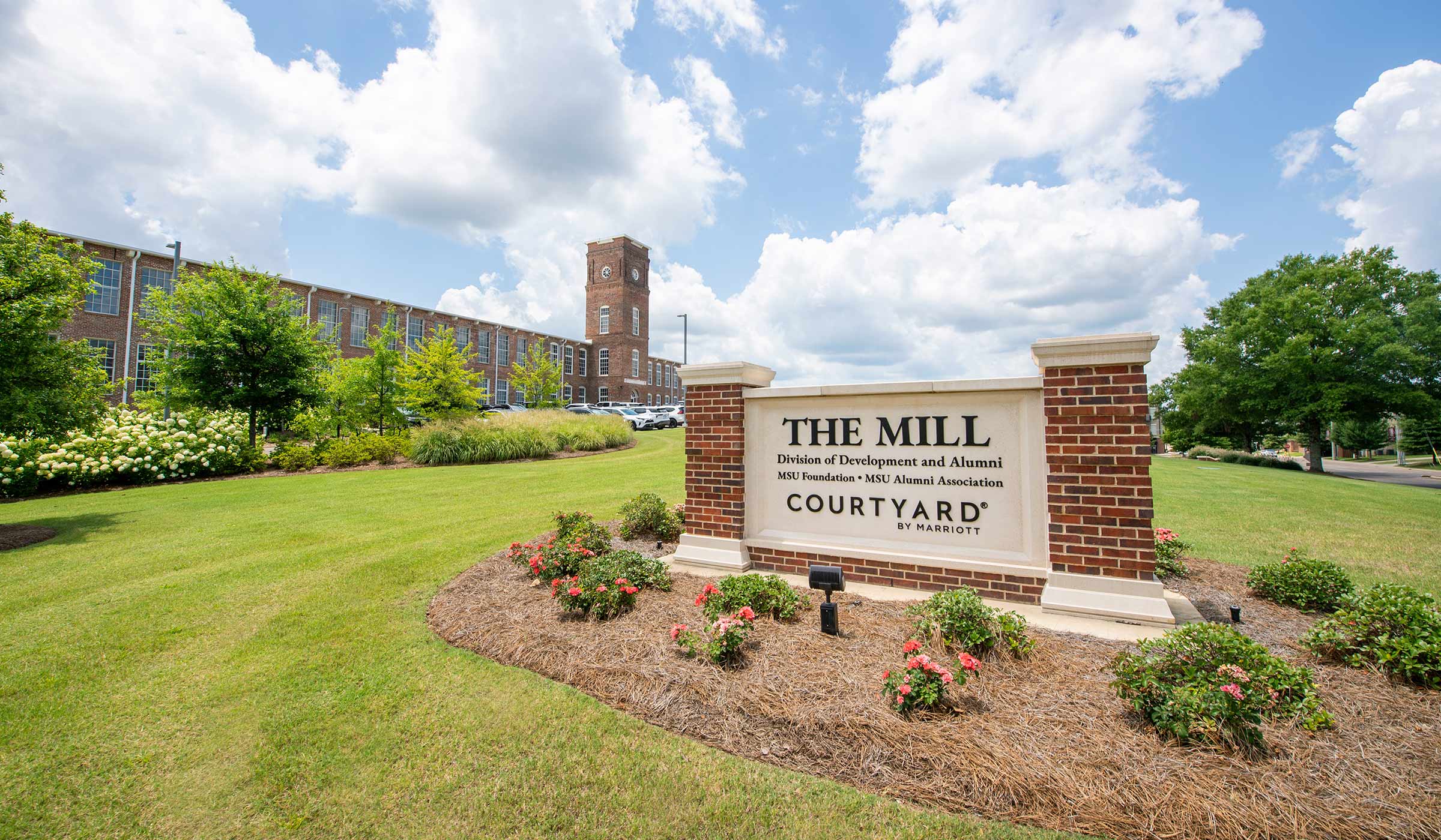 Brick and white sign in front of The Mill at MSU featuring the building surrounded by green grass and a blue sky.