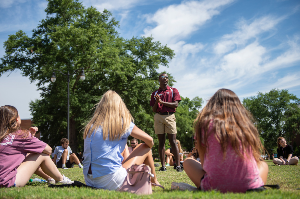 Student in maroon polo speaking in front of seated students on a beautiful blue sky day