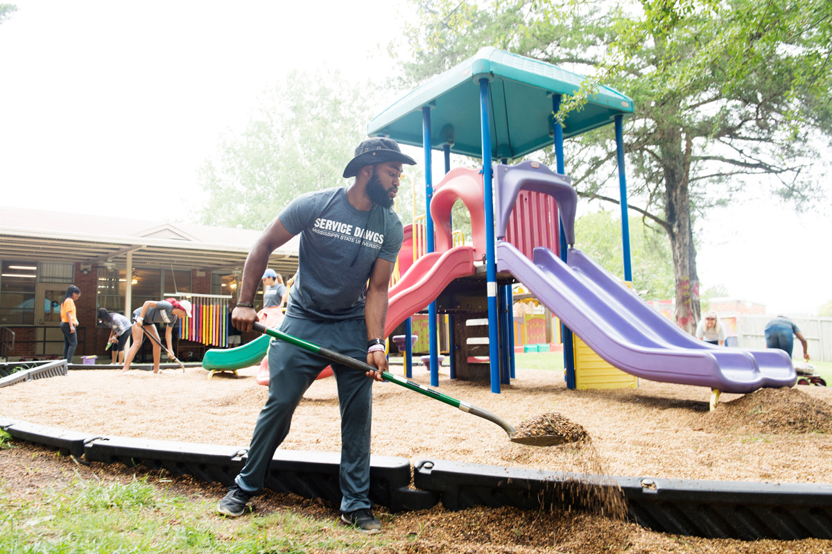 Student shovels gravel at Child Development Center playground with other Service Dawgs Day volunteers working in background.