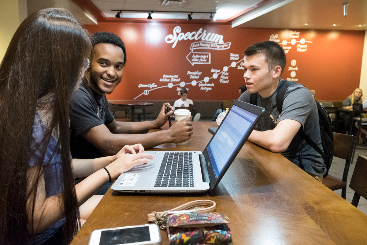 Three friends chat over coffee and a laptop at Starbucks table.