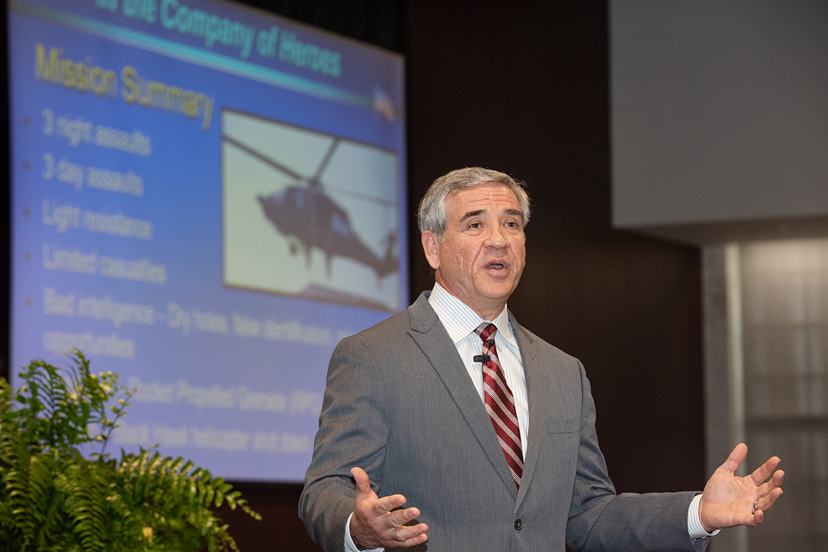 Man in gray suit speaking in front of projected image of helicopter and words