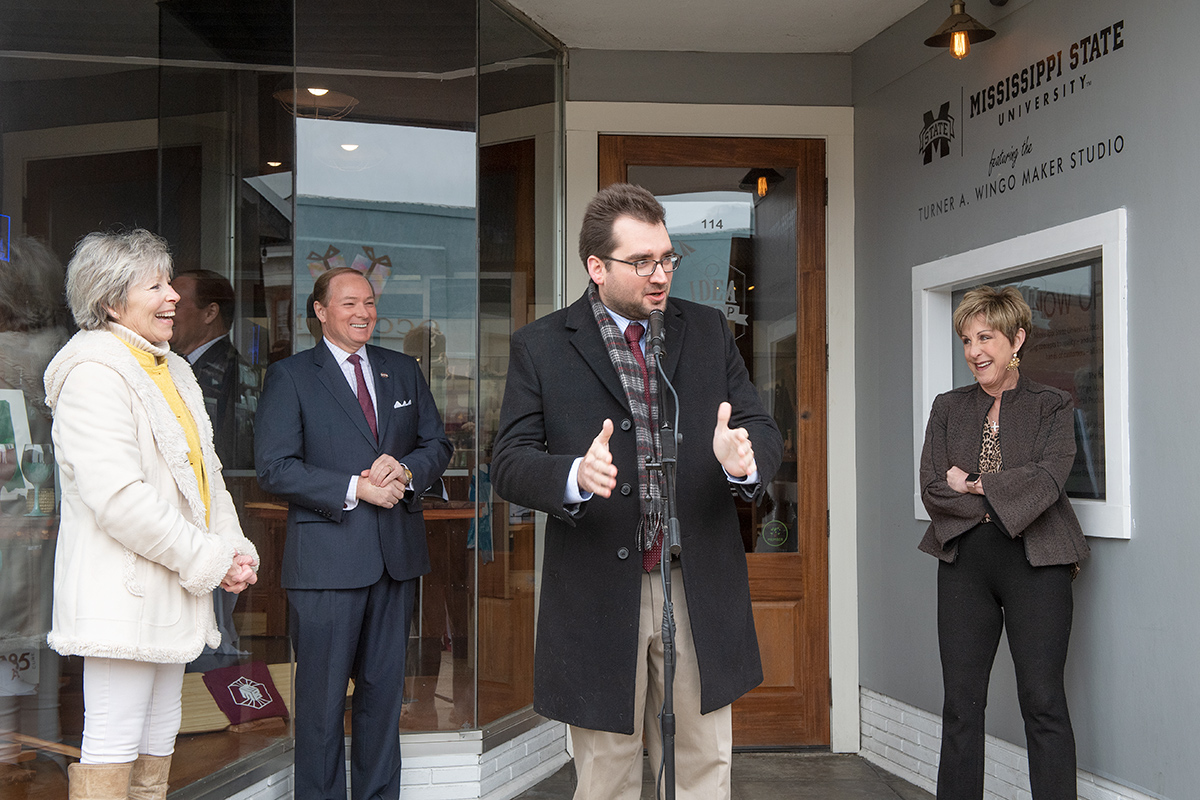 Man in suit and overcoat speaking at microphone with three people laughing at his joke