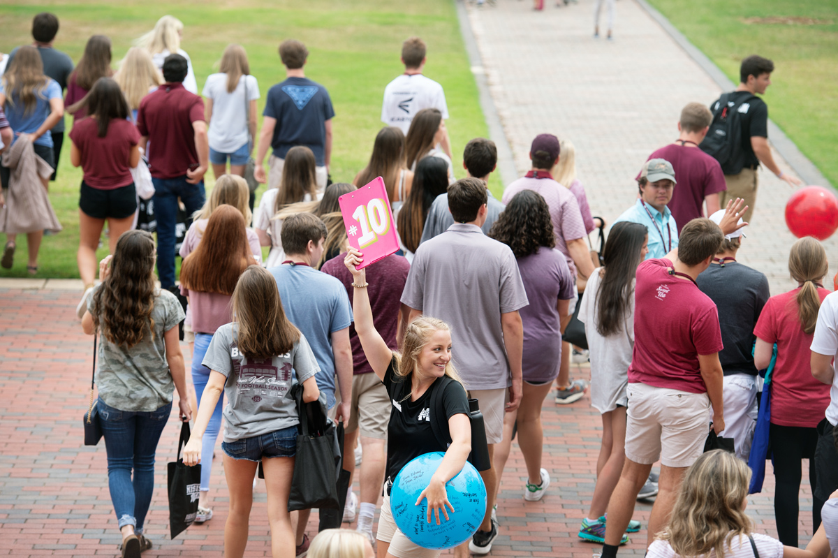 Female Orientation Leader holds up her number ten clipboard and ball on the Drill Field with Orientation attendees crowd behind.