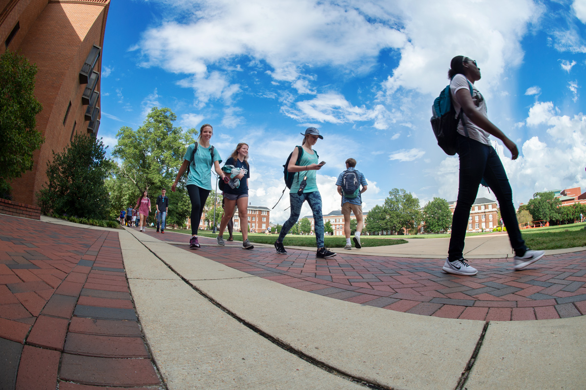 Students walk on sidewalk in front of McCool Hall, with Drill Field and clouds behind.