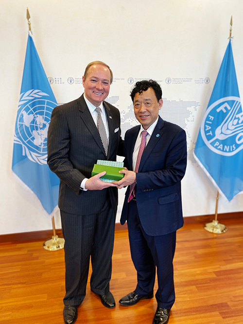 Two men stand together smiling at the camera with flags in the background.