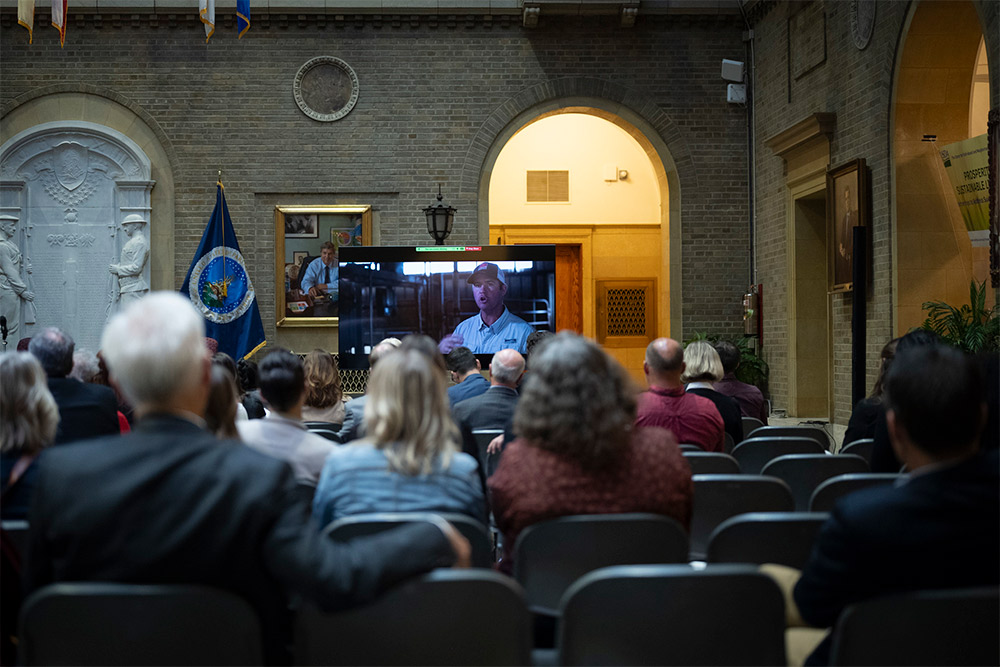 A group of people watching a large television screen with a farmer talking