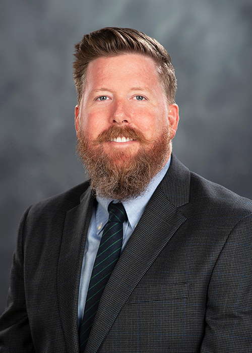 Studio portrait of Josh Foreman wearing a coat and tie in front of a gray background