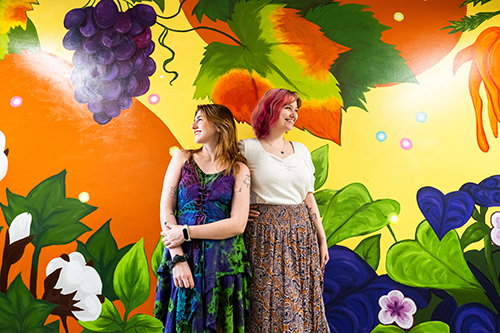 Senior Mississippi State art major Chloe Ruggiero of Covington, Louisiana, left, and spring 2024 graduate Abbey Temple of Ridgeland, stand in front of the 32-foot-long mural they created at MSU’s Hill Agriculture Science Building. The painting depicts select Mississippi crops and the diseases that plague them.