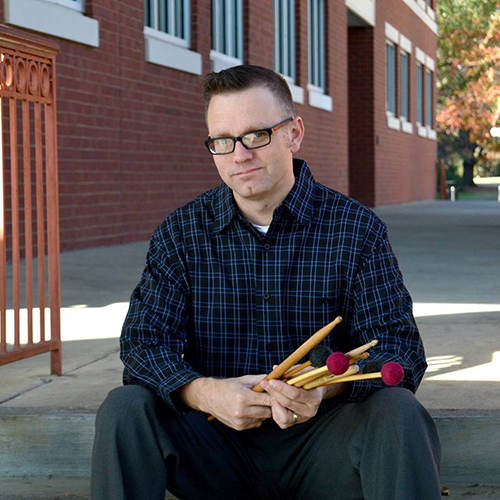 MSU Professor of Music Jason Baker sits outside on concrete steps while holding marimba mallets.