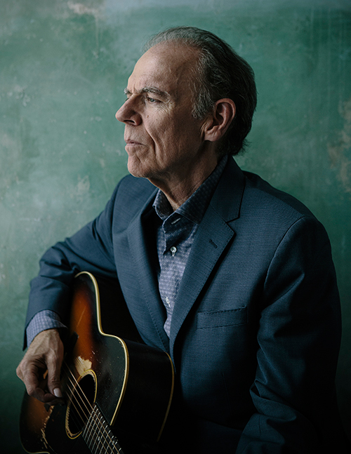 Singer-songwriter John Hiatt stares to his left while holding a guitar.