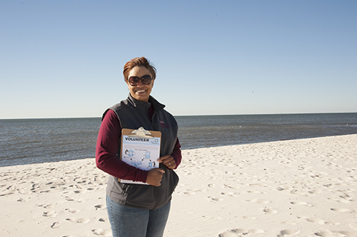 Image of a smiling lady wearing a maroon jacket and holding a clipboard on the beach with the ocean in the background