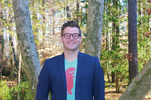 A man is pictured wearing glasses and a blazer and T-shirt with a wooded area in the background