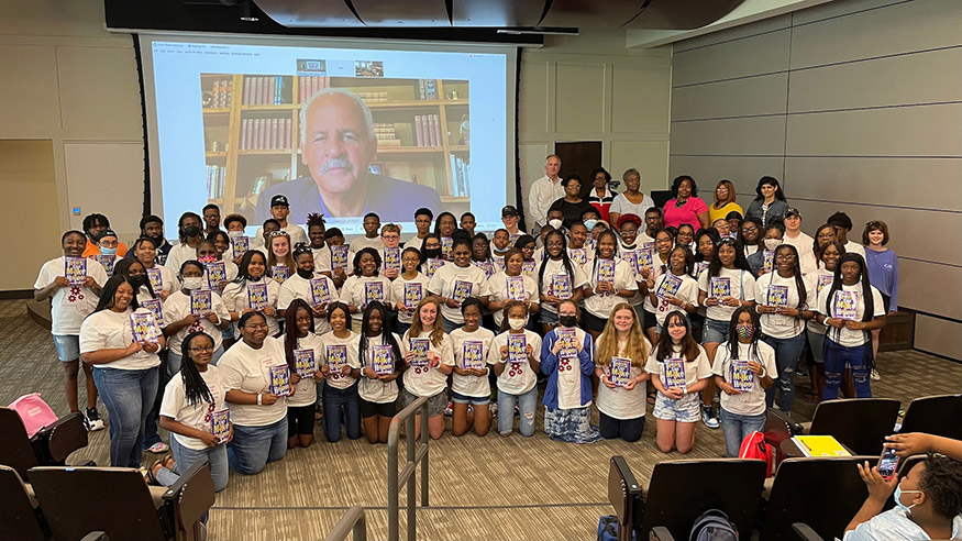 A large group of Mississippi students holding books, along with some teachers, stand in front of a large screen showing their virtual guest speaker, Stedman Graham.