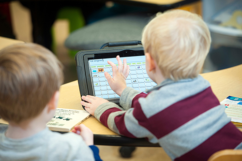 Two children, viewed from behind, look over a touch-screen computer.