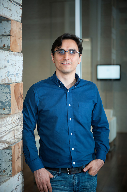 Farshid Vahedifard stands inside a building by a wooden wall wearing a blue shirt and jeans