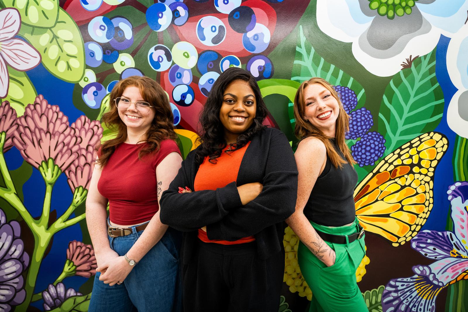 three women stand in front of a colorful mural