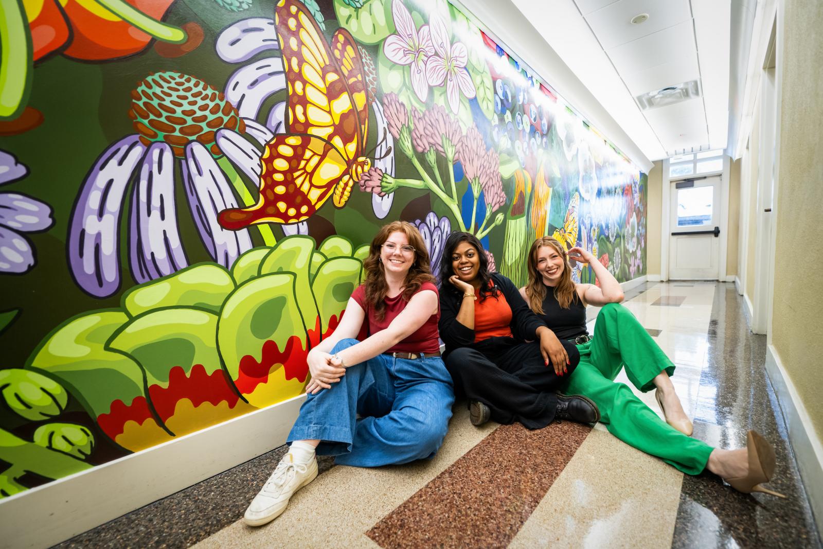 three women sit in hallway in front of colorful mural