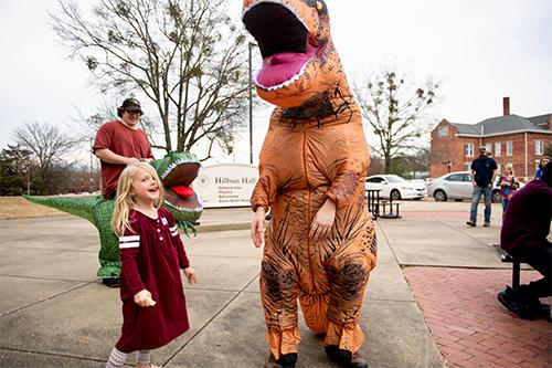 A young girl smiles at a nearby person in a dinosaur costume