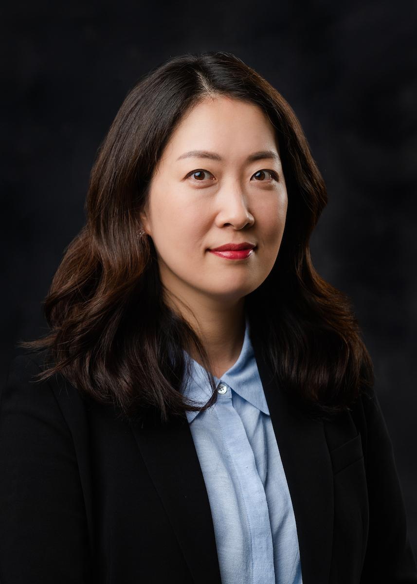headshot of woman in blue shirt and black blazer on dark background.