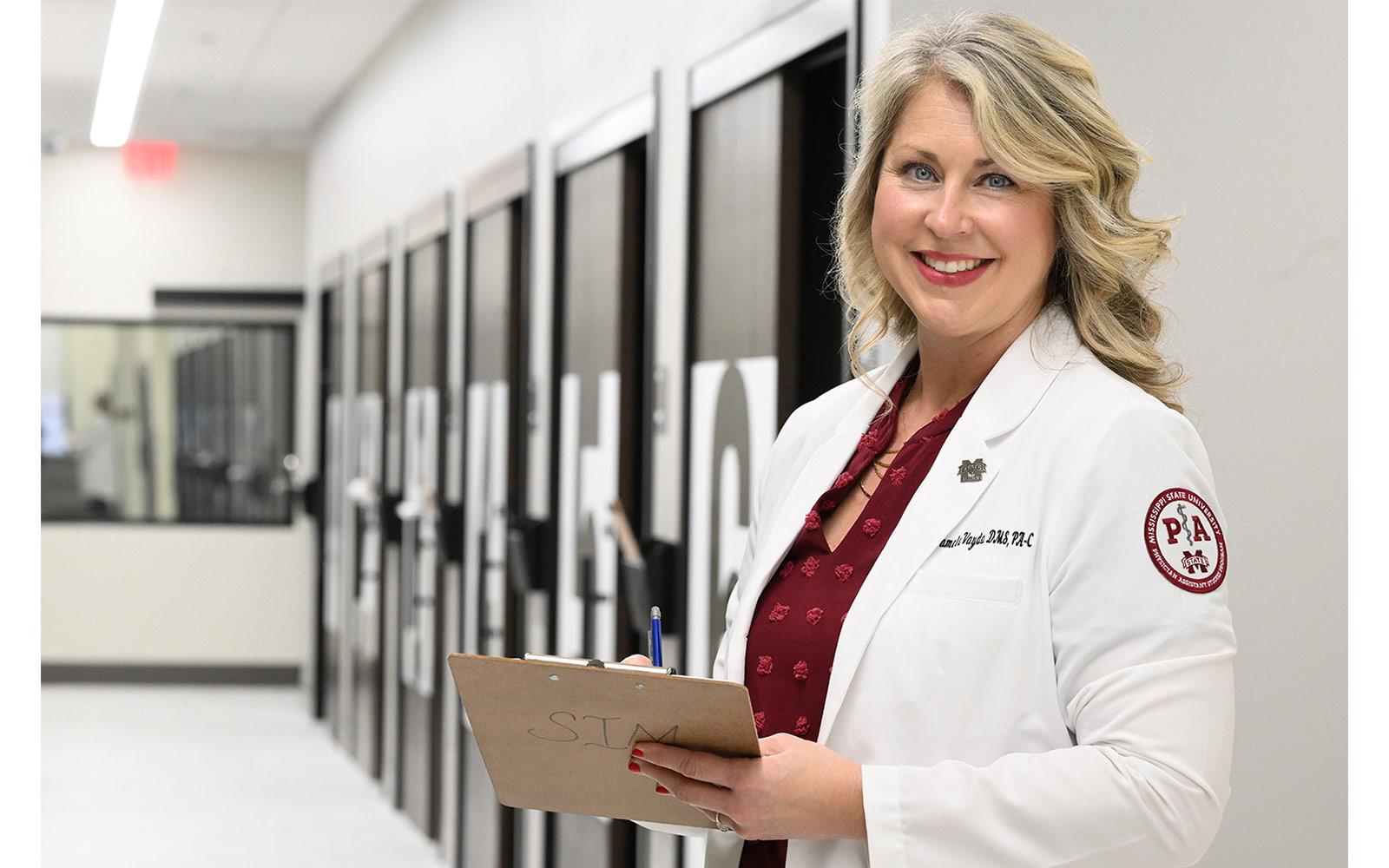 Pamela Vayda in her clinical white coat holding a clipboard at MSU-Meridian's Interprofessional Simulation facility.