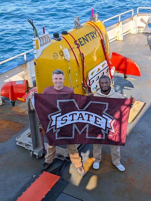Mississippi State Department of Geosciences Professor Adam Skarke, left, and Ramon Kalski, a Ph.D. student in earth and atmospheric sciences from Pascagoula, pose on the deck of a ship while holding a MSU flag.